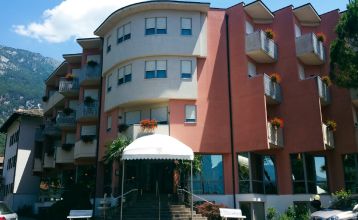 Luftaufnahme des Hotels Everest***, eines rosafarbenen mehrstöckigen Gebäudes mit renovierten Zimmern, umgeben von Grünanlagen, einer kleinen Kirche, Häusern, Gärten, bewaldeten Hügeln und nahe gelegenen Fußball- und Trainingsplätzen, die sich in die frie