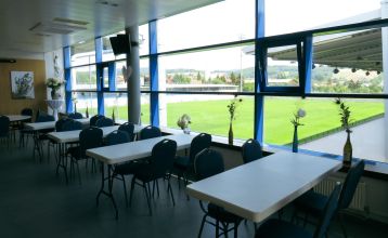 Der Gasthof & Gästehaus Trummer, ein hellblauer Gasthof mit rotem Dach, steht an einer von Bäumen gesäumten Straße nahe der Sportanlage Ritzing. Bei teilweise bewölktem Himmel sind parkende Autos und andere Häuser in der Nähe.