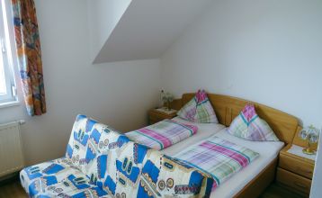 Der Gasthof & Gästehaus Trummer, ein hellblauer Gasthof mit rotem Dach, steht an einer von Bäumen gesäumten Straße nahe der Sportanlage Ritzing. Bei teilweise bewölktem Himmel sind parkende Autos und andere Häuser in der Nähe.