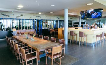 Der Gasthof & Gästehaus Trummer, ein hellblauer Gasthof mit rotem Dach, steht an einer von Bäumen gesäumten Straße nahe der Sportanlage Ritzing. Bei teilweise bewölktem Himmel sind parkende Autos und andere Häuser in der Nähe.