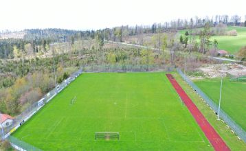 Luftaufnahme des Breitenberger Hofs*** mit Sonnenkollektoren und einem Wellnessbereich auf dem Dach, umgeben von geparkten Autos und Häusern in einer kleinen, hügeligen Stadt bei Passau. Auf dem Boden sind Schneeflecken zu sehen.