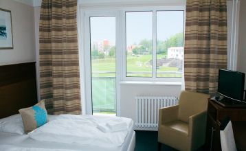 Das Hotel Marttel*** ist ein modernes weißes Gebäude mit vielen Fenstern in der Nähe des grünen Kunstrasen-Fußballplatzes, umgeben von Bäumen und Hügeln, mit Wohnhäusern im Hintergrund unter einem teilweise bewölkten Himmel, nahe der deutschen Grenze.