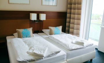 Das Hotel Marttel*** ist ein modernes weißes Gebäude mit vielen Fenstern in der Nähe des grünen Kunstrasen-Fußballplatzes, umgeben von Bäumen und Hügeln, mit Wohnhäusern im Hintergrund unter einem teilweise bewölkten Himmel, nahe der deutschen Grenze.