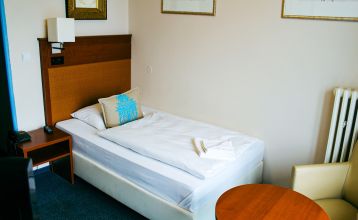 Das Hotel Marttel*** ist ein modernes weißes Gebäude mit vielen Fenstern in der Nähe des grünen Kunstrasen-Fußballplatzes, umgeben von Bäumen und Hügeln, mit Wohnhäusern im Hintergrund unter einem teilweise bewölkten Himmel, nahe der deutschen Grenze.