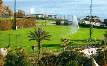 Luftfoto av Limak Arcadia Hotel, et livlig strandkompleks med bassenger, tennisbaner, hager, sandstrand med parasoller og brygge - ideelt for en fotballtreningsleir i Tyrkia. Omgitt av tette trær.