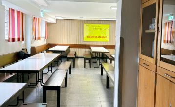 Das Gästehaus Nettersheim ist ein modernes, dreistöckiges Gebäude mit großen Fenstern, einer roten unteren Fassade, hölzernen Obergeschossen und einem kleinen Balkon, eingebettet in eine grüne, natürliche Umgebung mit Bäumen unter der Sonne.