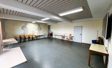 Das Gästehaus Nettersheim ist ein modernes, dreistöckiges Gebäude mit großen Fenstern, einer roten unteren Fassade, hölzernen Obergeschossen und einem kleinen Balkon, eingebettet in eine grüne, natürliche Umgebung mit Bäumen unter der Sonne.