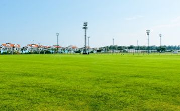 Luftfoto av Limak Arcadia Hotel med bassenger, hager, idrettsbaner og strandhytter - ideelt for fotballtreningsleiren i Tyrkia - omgitt av palmer og fjell under en klar himmel.