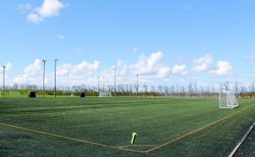 Eine Reihe kleiner, gelber Holzhütten mit roten Dächern säumt einen geschwungenen Weg im Sportzentrum Skaerbaek, umgeben von grünem Gras und Bäumen unter einem hellen, teilweise bewölkten Himmel.