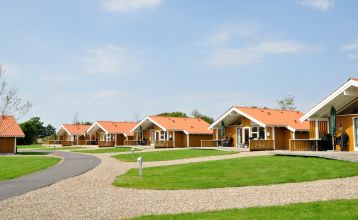 Eine Reihe kleiner, gelber Holzhütten mit roten Dächern säumt einen geschwungenen Weg im Sportzentrum Skaerbaek, umgeben von grünem Gras und Bäumen unter einem hellen, teilweise bewölkten Himmel.