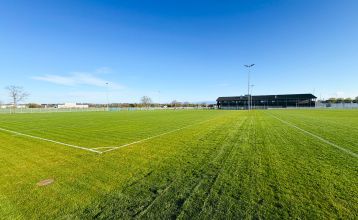 Das Egger Stüberl ist ein weißes, zweistöckiges Gebäude mit rotem Ziegeldach und Balkonen unter strahlend blauem Himmel. Perfekt für Besucher oder Trainingslager in der Nähe lokaler Fußballplätze, bietet es Sitzgelegenheiten im Freien und Pflanzen zur Beg