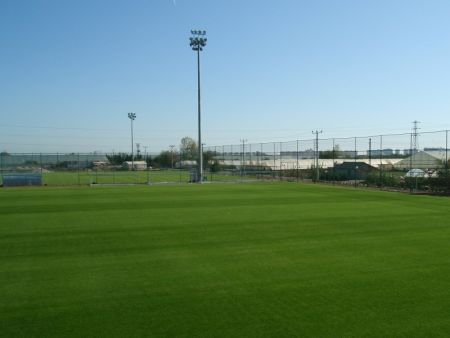 Titanic Deluxe Lara Footballcenter Platz A zeigt ein gepflegtes, grünes Spielfeld unter freiem Himmel, umgeben von hohen Flutlichtern und einem Drahtzaun, mit Gebäuden und anderen Feldern im Hintergrund.