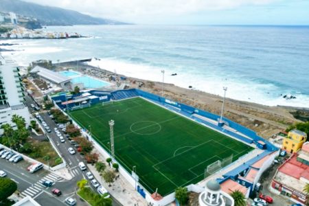 Luftfoto av ElPenon-kunstgressbanene, en fotballbane som ligger rett ved stranden med tribuner og et svømmebasseng i nærheten, omgitt av bygninger og gater under en overskyet himmel.