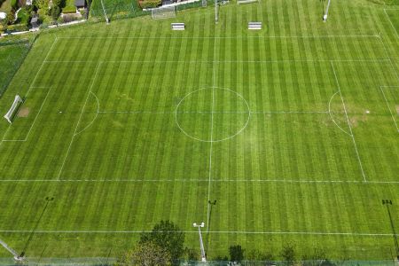 Luftfoto av Eisenstadts naturgressbane, en velholdt fotballbane med markerte hvite linjer, grønnstripet gress, benker, belysning og trær i kanten.