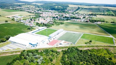 Luftaufnahme des Trainingsplatzes Vinea Resort - ein Sportkomplex mit einem Stadion, grünen Feldern und einer kleinen Stadt, Ackerland und bewaldeten Hügeln in der ländlichen Landschaft unter einem klaren Himmel.