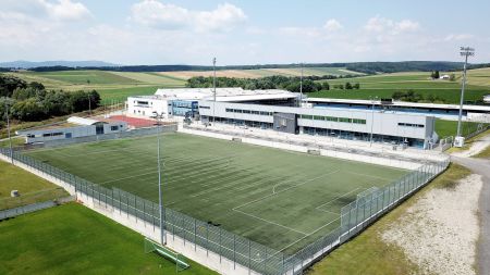 Luftaufnahme des Kunstrasenplatzes Vinea Resort, eines leeren Fußballplatzes mit Kunstrasen, eingezäunt und mit Flutlicht beleuchtet, neben modernen Sportanlagen und Grünflächen unter einem teilweise bewölkten Himmel.