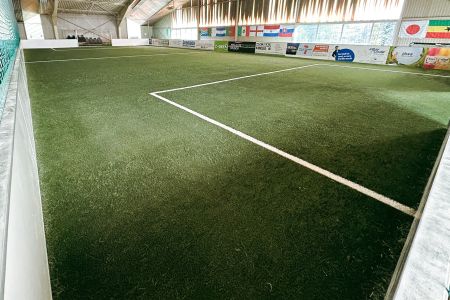 Der FunCourt Wiesloch verfügt über ein Hallenfußballfeld mit Kunstrasen, grünem Netz auf der linken Seite, weißen Begrenzungslinien und Länderflaggen und Werbung an der gegenüberliegenden Wand unter einer hohen, gut beleuchteten Decke.