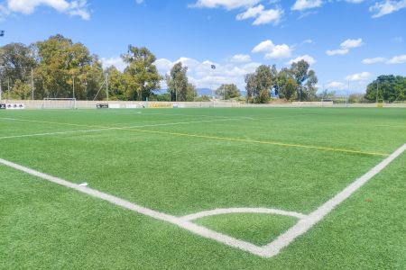 På en klar, solrik dag er Campo de Futbol Guadalmar en grønn kunstgressbane med hvite og gule feltmarkeringer. Trær danner bakgrunnen under en blå himmel med spredte skyer. Det er ingen spillere på banen.