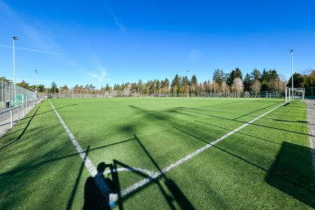 En solrik dag på Kunstrasenplatz Gärtringen, en tom utendørs fotballbane med knallgrønt kunstgress, hvite banemarkeringer og trær i bakgrunnen. Skygger fra gjerder og utstyr kan ses på banen.