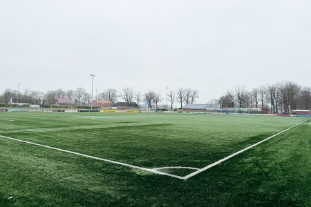 Vidvinkelbilde av den tomme, grønne fotballbanen til SV Grol Kunstrasen 2 på en overskyet dag, med hvite banelinjer og trær, bygninger og gjerder i bakgrunnen.