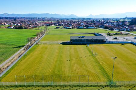 Luftaufnahme von Schechen Trainingsplatz 2, einem grünen Fußballplatz neben einem Gebäude, eingezäunt und begrenzt von Feldern und einer kleinen Stadt, mit Bergen in der Ferne unter einem klaren blauen Himmel.