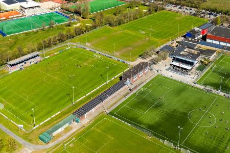 Luftaufnahme des Sportkomplexes Naturrasen 1 Wassenaar mit mehreren grünen Fußballplätzen, von denen einige genutzt werden und andere leer sind, umgeben von Gebäuden, Parkplätzen, Straßen und Bäumen.