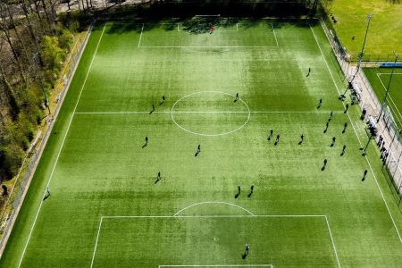 Die Luftaufnahme von Kunstrasen 2 Wassennaar zeigt die Spieler auf dem grünen Fußballfeld, einige in der Nähe des Mittelkreises, andere rechts, mit Bäumen und einem weiteren Feld in der Nähe.