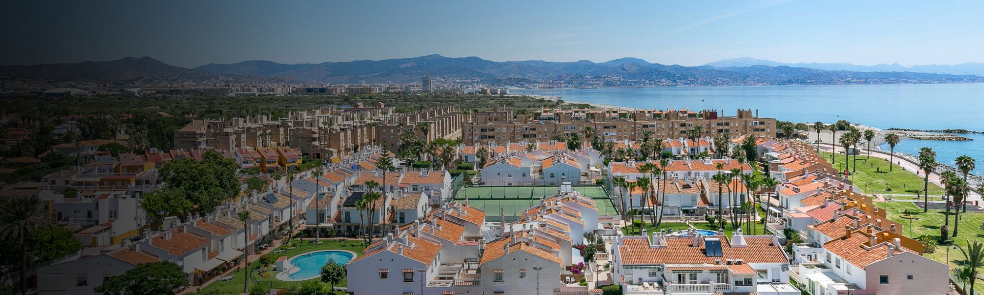 Panoramaaufnahme einer gro&szlig;en Fu&szlig;balltrainingsanlage in Spanien, bestehend aus mehreren Naturrasen- und Kunstrasenpl&auml;tzen, umgeben von landwirtschaftlichen Feldern, Olivenhainen und einer K&uuml;stenstadt mit dem Mittelmeer im Hintergrund.