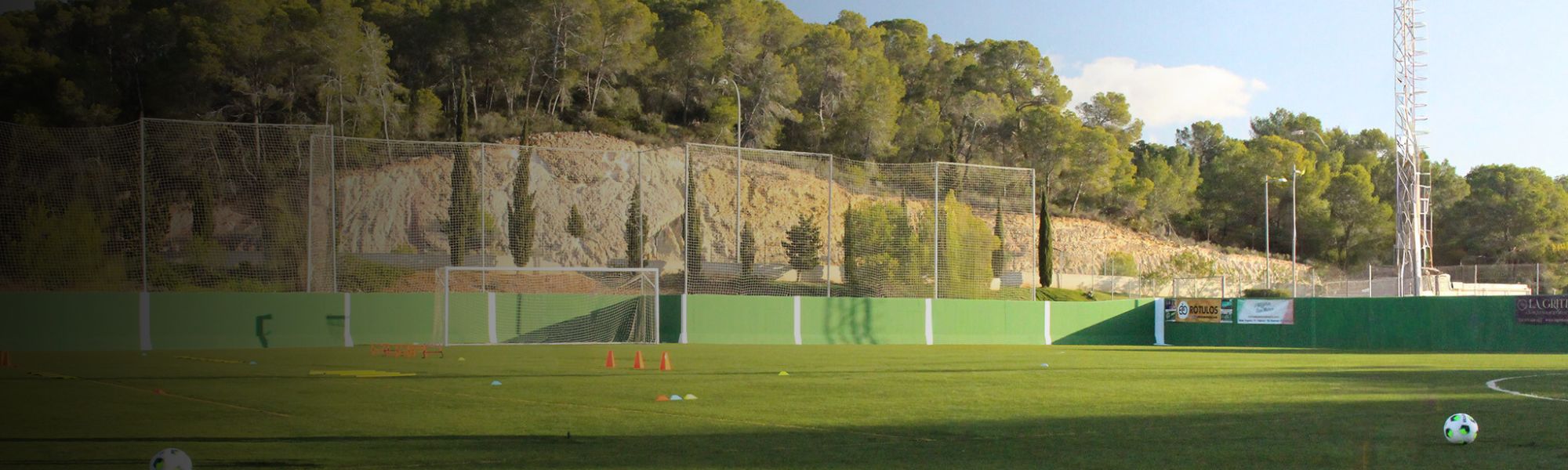 Kunstrasen-Fu&szlig;ballplatz auf Sardinien mit markierten Spielfeldlinien und Umz&auml;unung, im Hintergrund Berge und Geb&auml;ude.