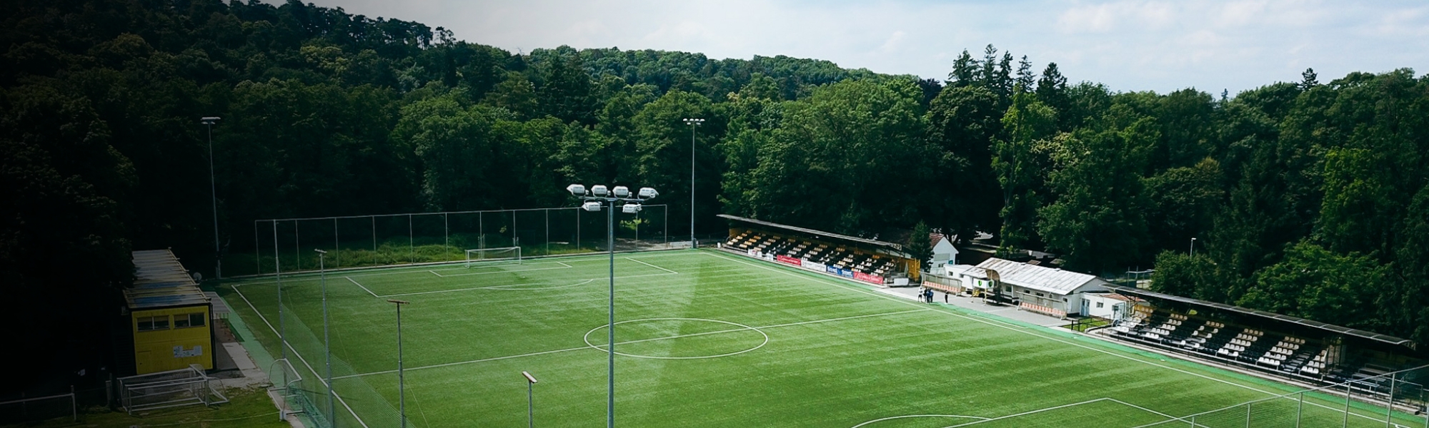 Fu&szlig;balltrainingsplatz umgeben von dichten W&auml;ldern mit einer kleinen Trib&uuml;ne und Flutlichtmasten.