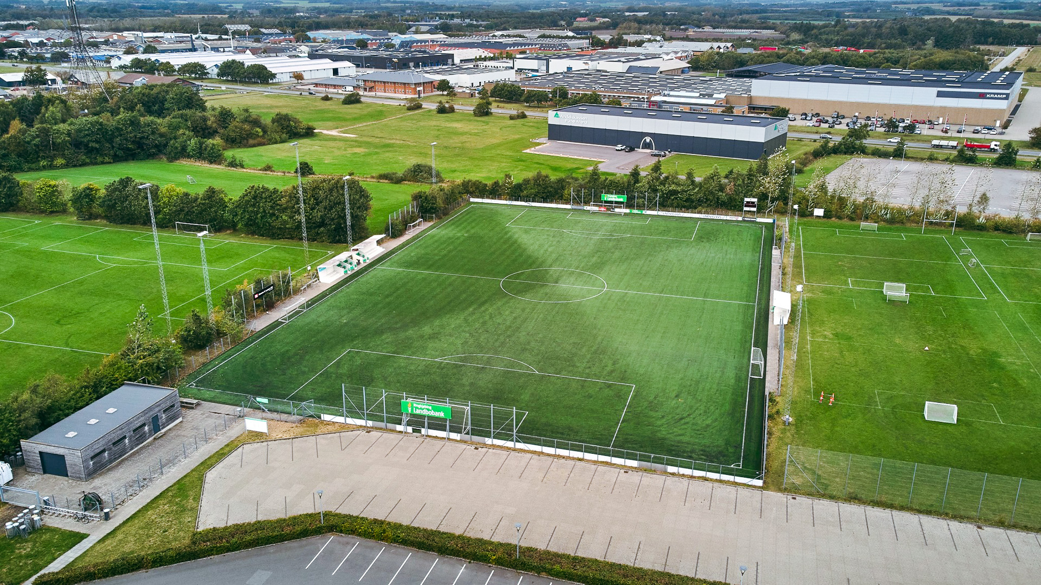 Luftaufnahme einer modernen Fu&szlig;balltrainingsanlage in D&auml;nemark mit einem gro&szlig;en Kunstrasenplatz, mehreren Naturrasenpl&auml;tzen, Flutlichtanlagen und angrenzenden Industrie- und Gewerbegeb&auml;uden im Hintergrund.