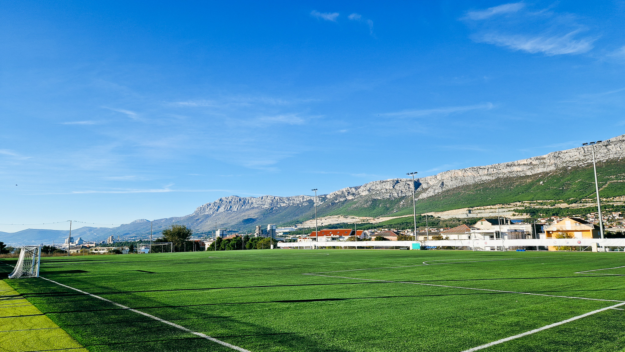 Sportanlage in Dalmatien mit einem Fu&szlig;ballfeld, Flutlichtmasten und Blick auf eine malerische Bergkette sowie Wohnh&auml;user im Hintergrund.