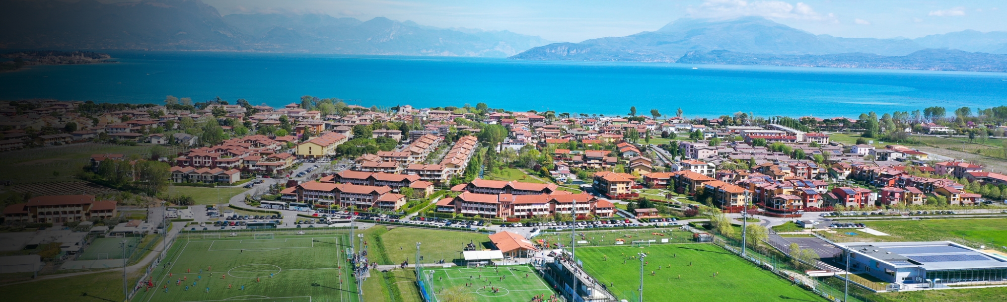 Panoramaaufnahme einer Sportanlage mit mehreren Fu&szlig;ballpl&auml;tzen am Gardasee, eingerahmt von Weinbergen, Wohnh&auml;usern und dem See im Hintergrund.