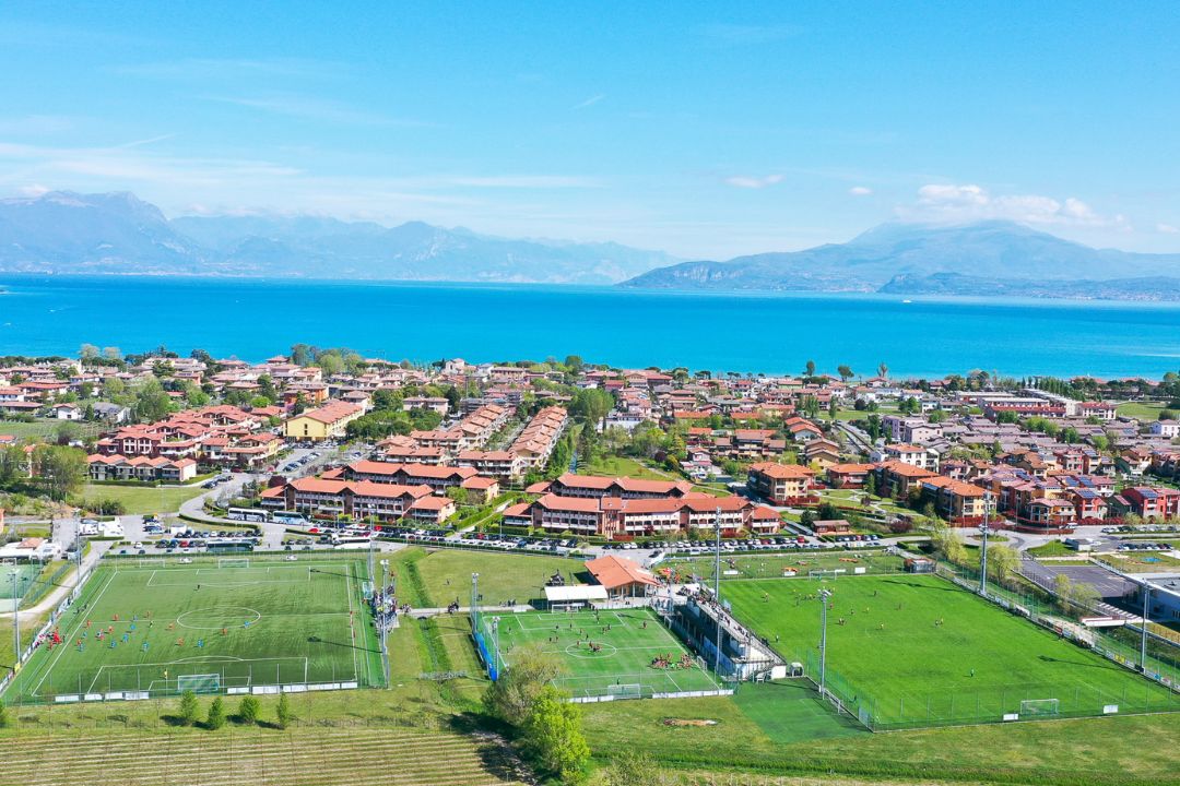 Luftaufnahme einer Fu&szlig;balltrainingsanlage am Gardasee, umgeben von roten Wohngeb&auml;uden, mit dem blauen See und Bergen im Hintergrund.