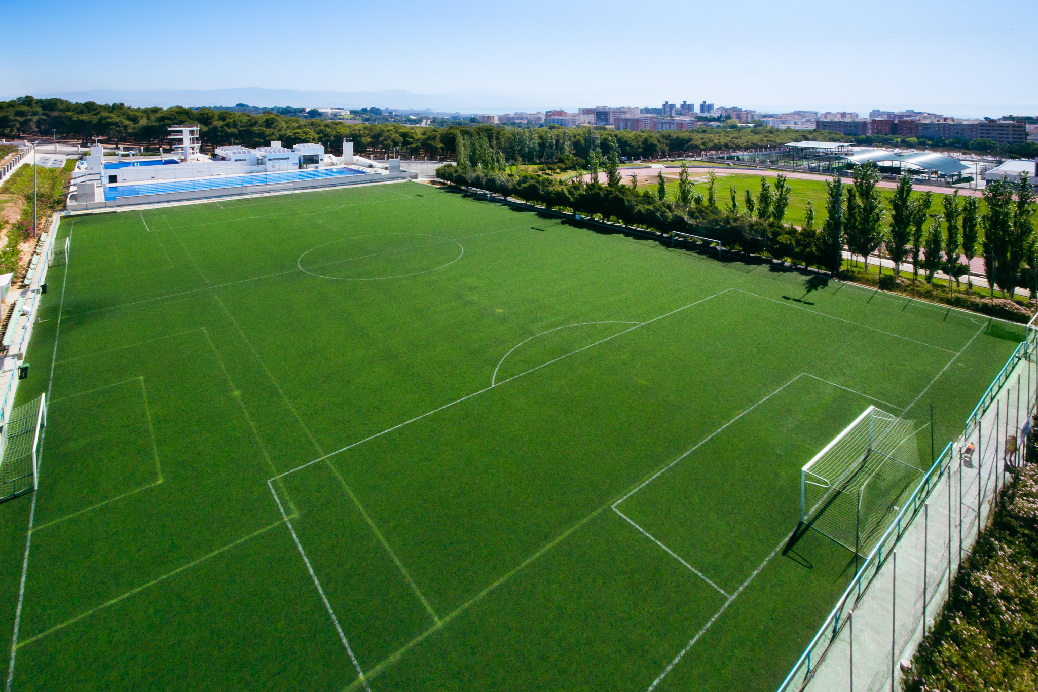 Fu&szlig;balltrainingsplatz auf Sardinien mit gr&uuml;nem Kunstrasen, Tor, B&auml;nken und Flutlichtmasten, umgeben von Netzen und Berglandschaft.