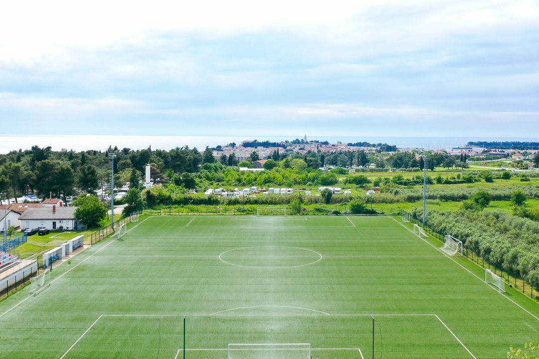  Fu&szlig;ballplatz in Istrien mit Kunstrasen, umgeben von gr&uuml;ner Natur und Olivenb&auml;umen, im Hintergrund das Meer und die Stadt Poreč.