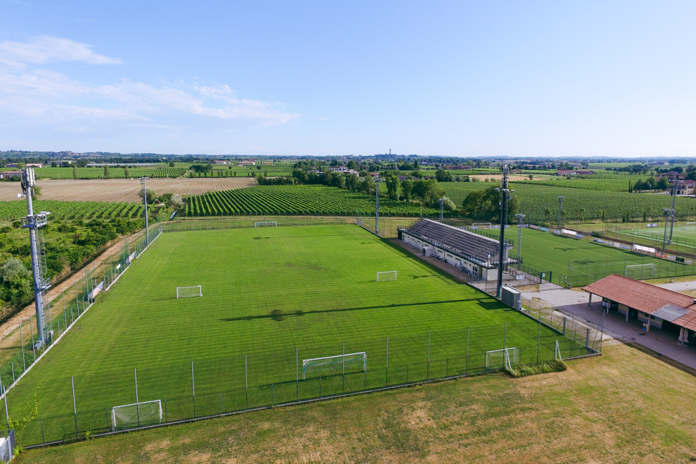 Luftaufnahme einer Sportanlage mit mehreren Fu&szlig;ballpl&auml;tzen, umgeben von einer italienischen Kleinstadt am Ufer eines Sees, mit Bergen im Hintergrund.
