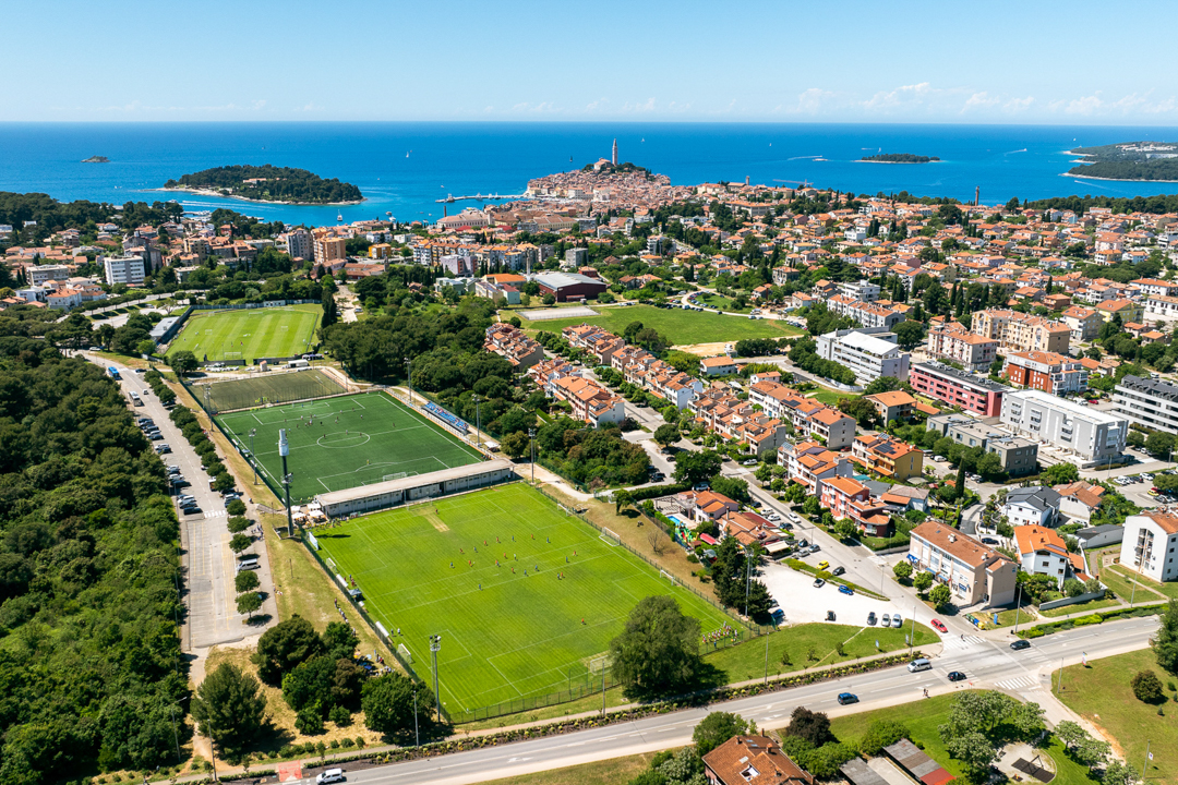 Luftaufnahme einer Fu&szlig;balltrainingsanlage in Kroatien mit mehreren Rasen- und Kunstrasenpl&auml;tzen, Flutlichtmasten, umgeben von Wohnh&auml;usern und Gr&uuml;nfl&auml;chen, im Hintergrund eine kleine Stadt und die Adria.