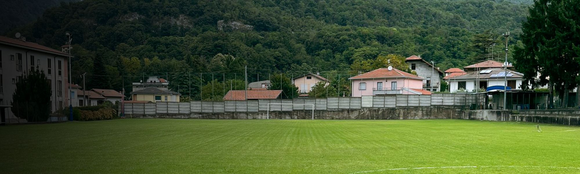 Kunstrasen-Fu&szlig;ballplatz auf Sardinien mit markierten Spielfeldlinien und Umz&auml;unung, im Hintergrund Berge und Geb&auml;ude.