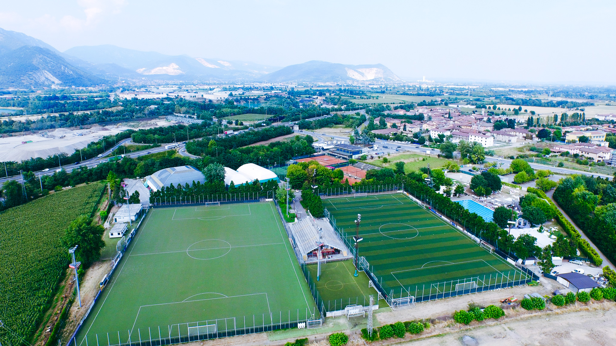 Weitl&auml;ufiger Blick auf eine Trainingsanlage in der Lombardei mit Fu&szlig;ballpl&auml;tzen, Tennisanlagen und einer Schwimmhalle, eingebettet in eine gr&uuml;ne Landschaft mit Bergen und Feldern im Hintergrund.