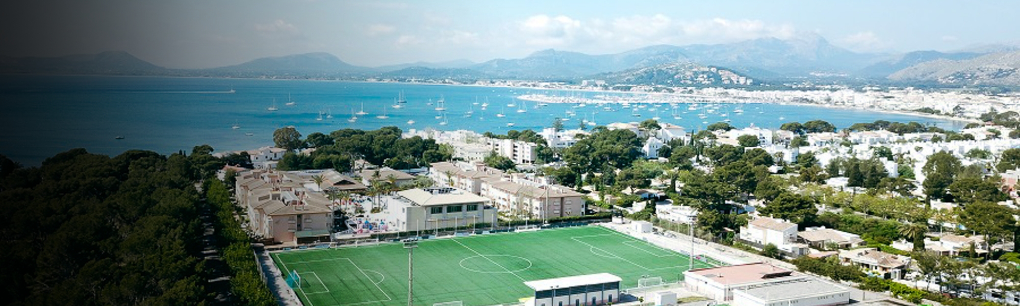 Panoramaaufnahme - Ein direkt am Meer gelegener Fu&szlig;ballplatz mit traumhafter Kulisse, Bergen und Segelbooten im Hintergrund.