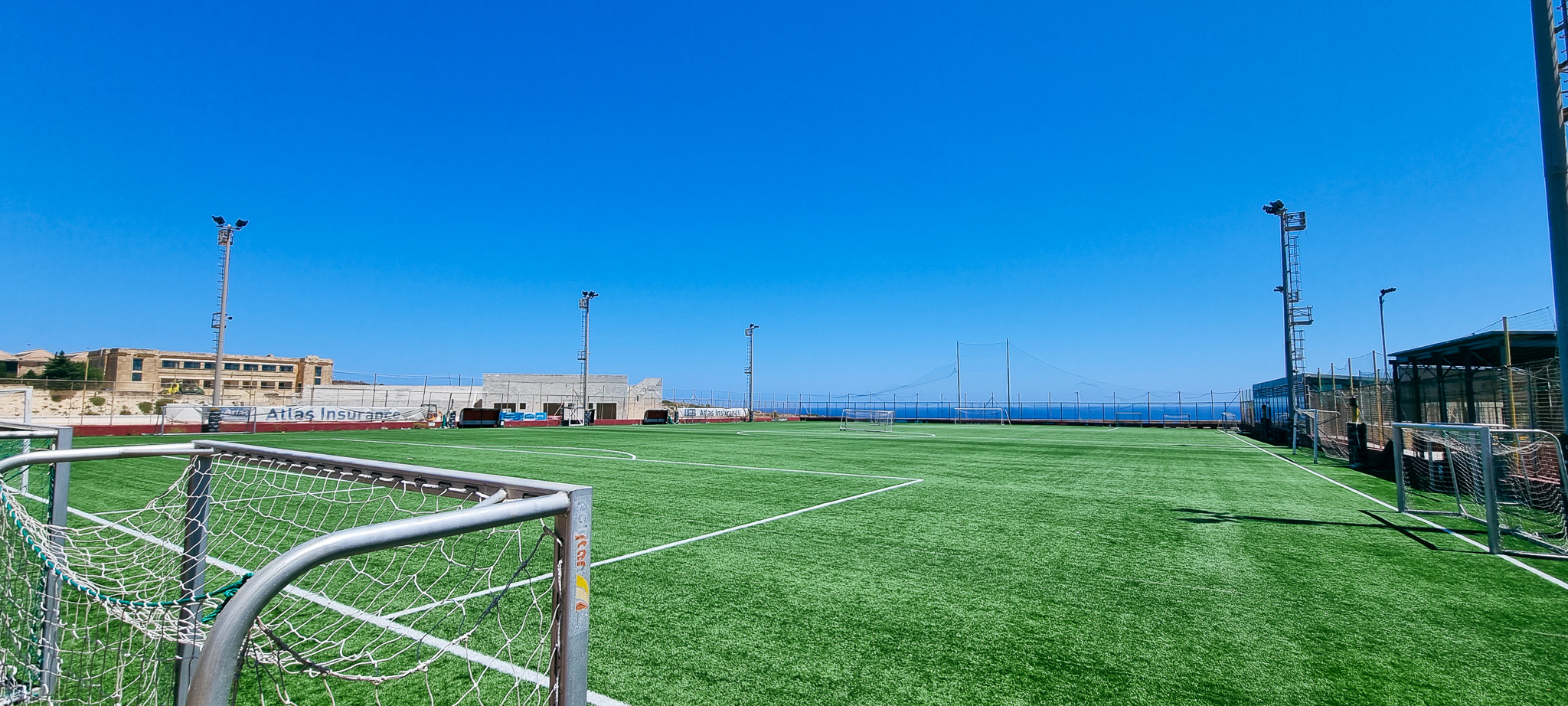 Fu&szlig;ballplatz mit Kunstrasen auf Malta, umgeben von Flutlichtmasten, mit Blick auf das Meer und einem klaren blauen Himmel im Hintergrund.