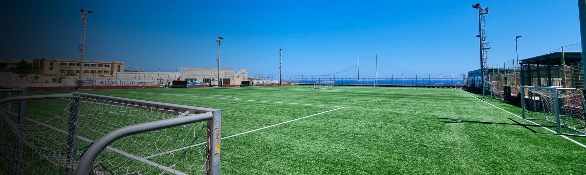 Fu&szlig;ballplatz mit Kunstrasen auf Malta, umgeben von Flutlichtmasten, mit Blick auf das Meer und einem klaren blauen Himmel im Hintergrund.