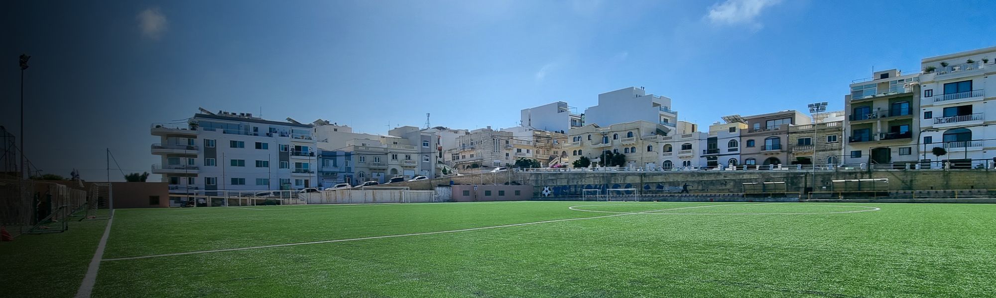 Panoramaaufnahme des Fu&szlig;ballplatzes in Mellieħa mit angrenzenden Wohnh&auml;usern &ndash; perfekte Trainingsbedingungen in einer sonnigen K&uuml;stenstadt.
