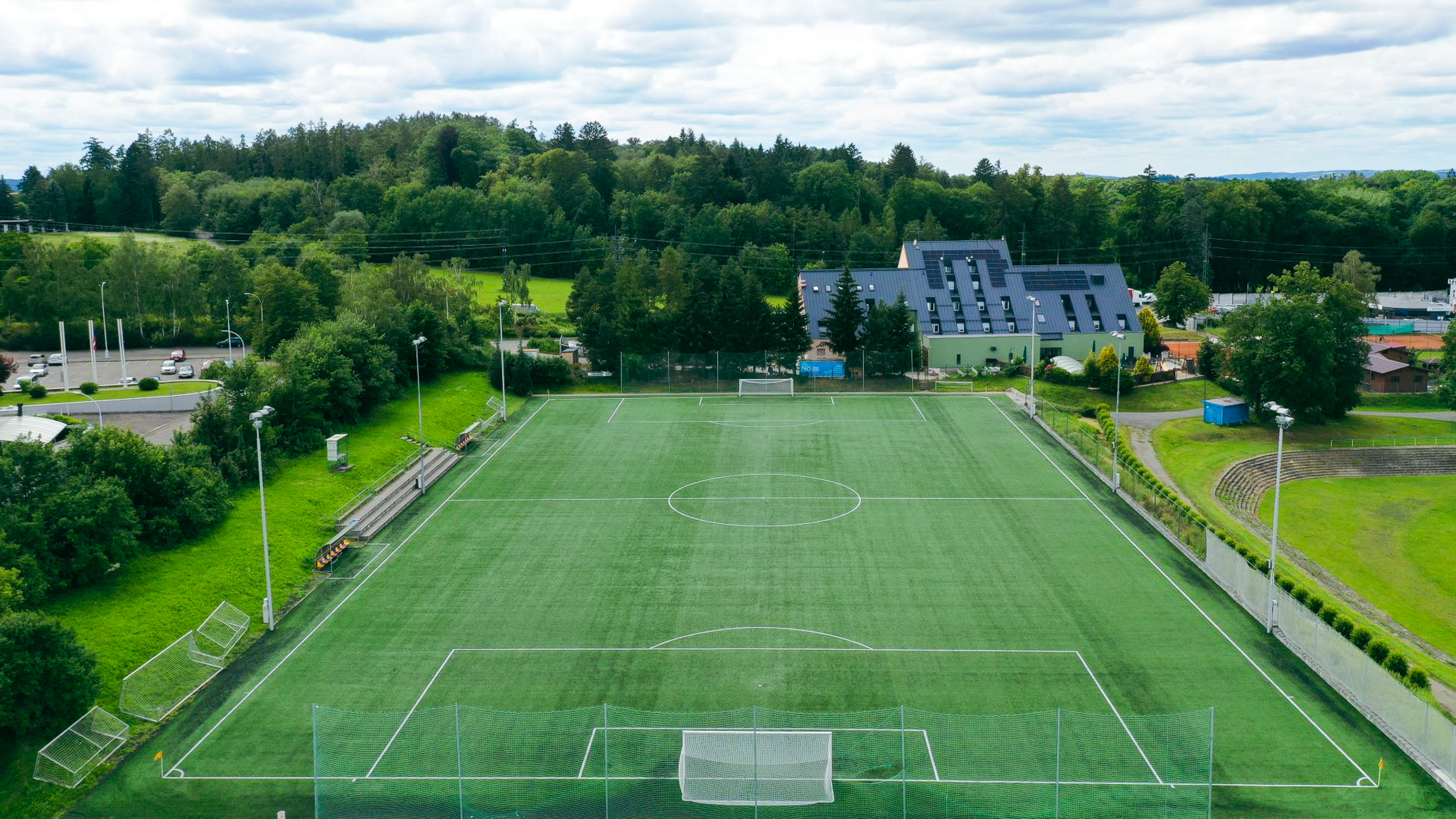 Fu&szlig;ballplatz mit Kunstrasen in einer idyllischen Umgebung, angrenzend an ein modernes Geb&auml;ude.