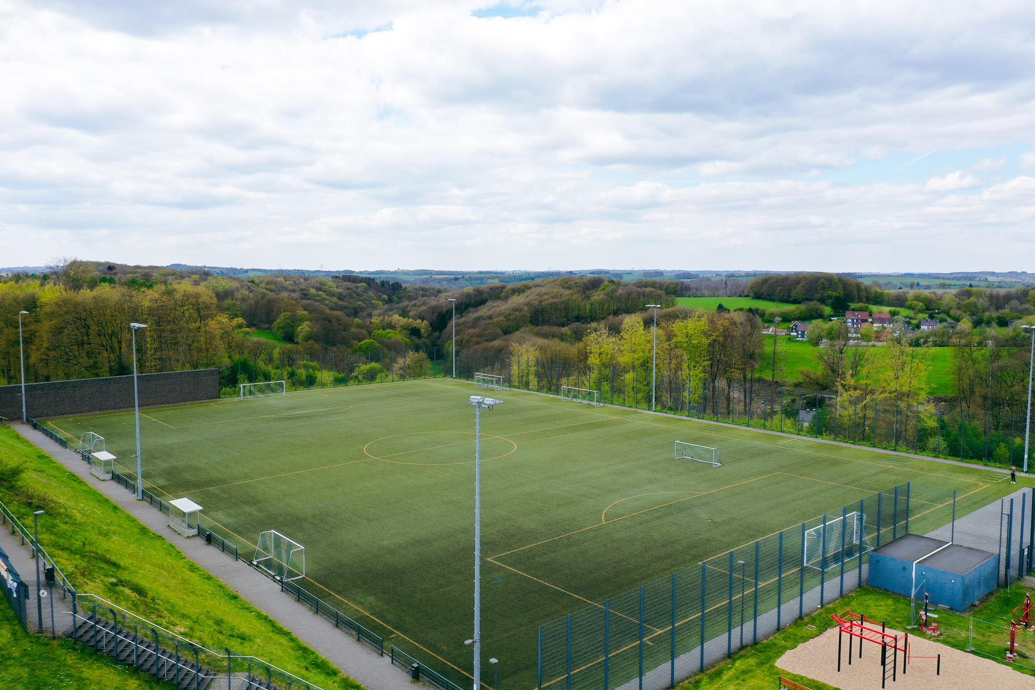 Luftaufnahme eines modernen Kunstrasen-Fu&szlig;ballplatzes in Nordrhein-Westfalen, umgeben von gr&uuml;nen H&uuml;geln und l&auml;ndlicher Landschaft.