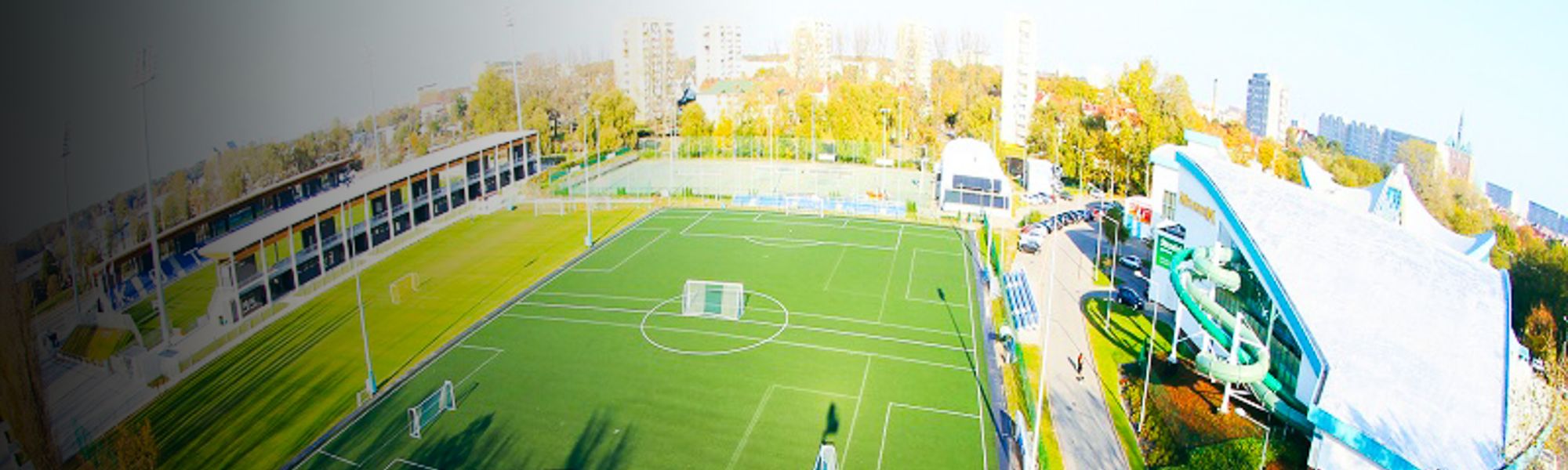 Panoramaansicht eines Fu&szlig;balltrainingszentrums in Polen mit Kunstrasenplatz, Trib&uuml;ne und benachbartem Erlebnisbad mit Rutsche.