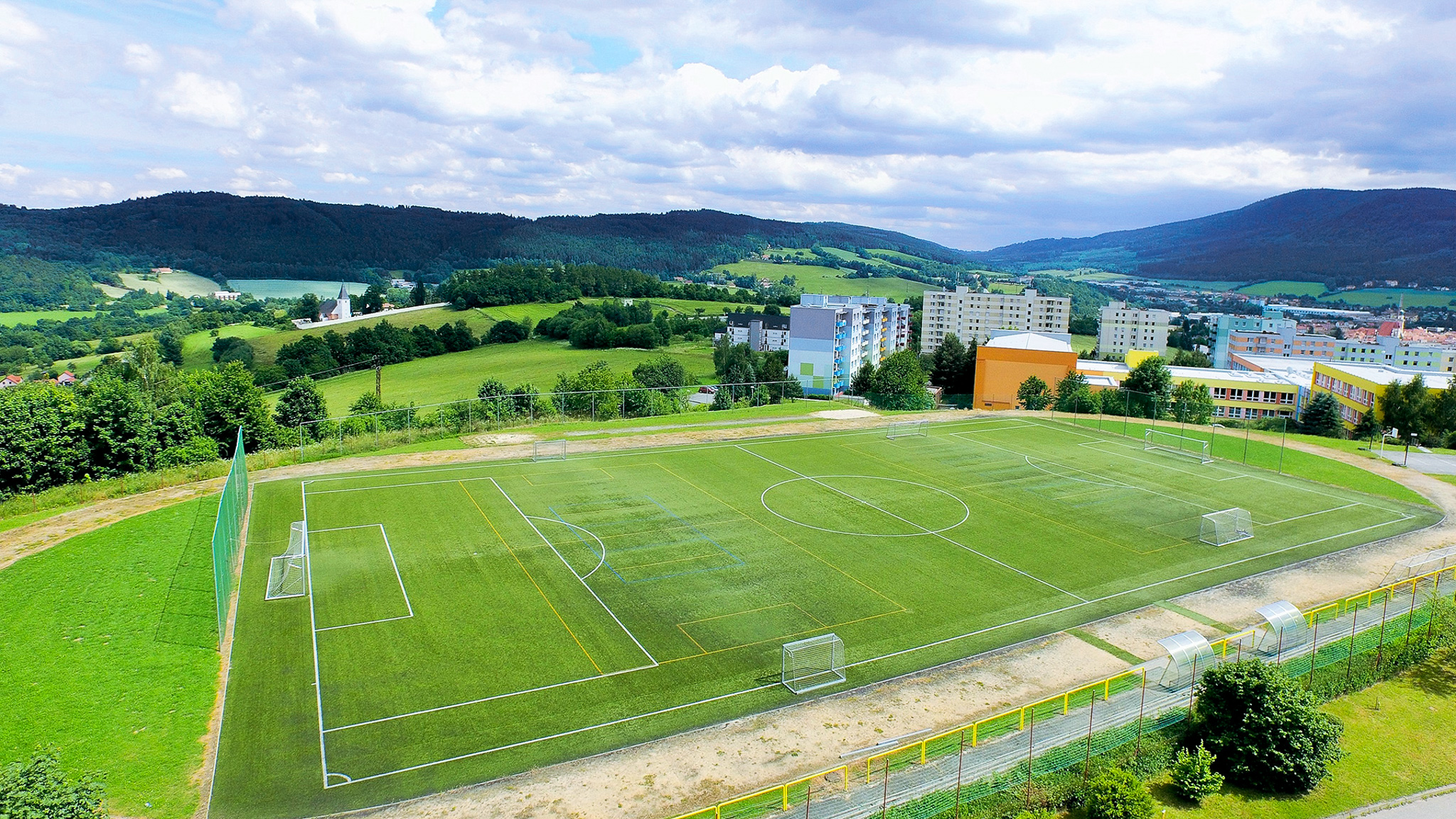 Fu&szlig;ballfeld mit Kunstrasen in einer malerischen H&uuml;gellandschaft, umgeben von Wohnh&auml;usern.