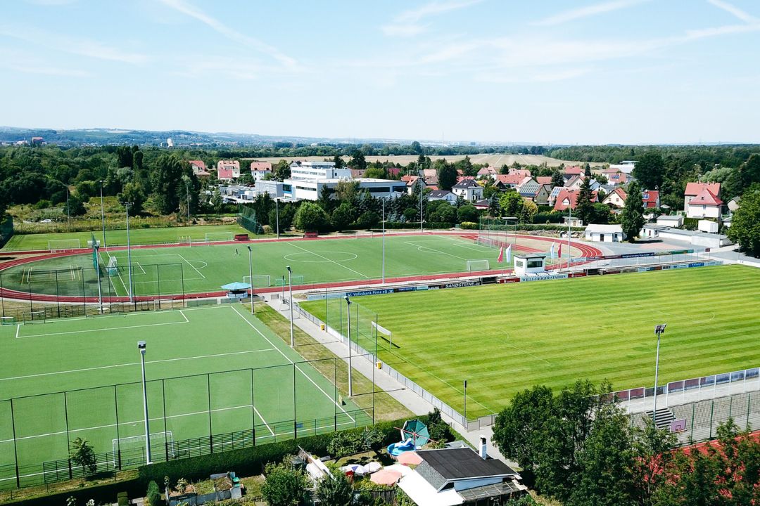 Sportanlage mit Fu&szlig;ballpl&auml;tzen, Laufbahn und Stadion, umgeben von Wohnh&auml;usern und gr&uuml;ner Landschaft.