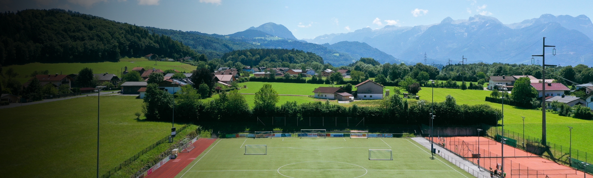 Fu&szlig;balltrainingsanlage in Salzburg mit Kunstrasenplatz, roten Laufbahnen und Tennispl&auml;tzen, eingebettet in eine idyllische Alpenlandschaft.